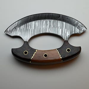 May include: A crescent-shaped ulu knife featuring a patterned Damascus steel blade. The handle is constructed from two dark wood sections and one lighter wood center piece, secured with three brass rivets and brass spacers.