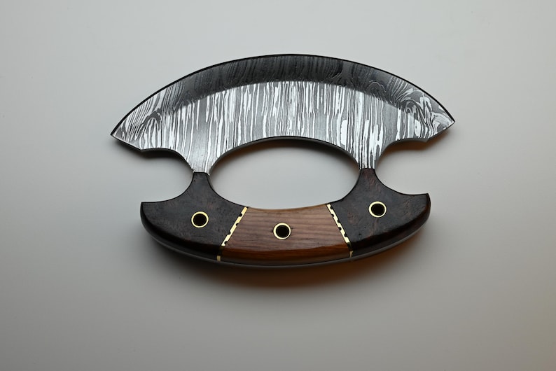 May include: A crescent-shaped ulu knife featuring a patterned Damascus steel blade. The handle is constructed from two dark wood sections and one lighter wood center piece, secured with three brass rivets and brass spacers.