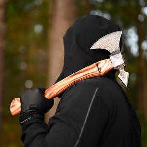 Könnte beinhalten: Eine Person in einem schwarzen Kapuzenpullover hält eine handgefertigte Axt mit einem Holzstiel. Die Axt hat eine Klinge aus Damaststahl mit einem einzigartigen Muster.