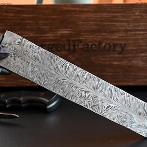 May include: A long, rectangular, patterned metal blade with a feather-like design is held by a gloved hand. A partially visible knife with a black handle and a wooden box with the text "ForgedFactory" are in the background.