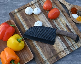 May include: A black cleaver knife with a wooden handle sits on a wooden cutting board with red and yellow bell peppers, garlic, tomatoes, and spices in a wooden bowl.