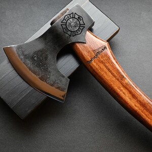 May include: Axe with a wooden handle and a metal head, resting on a dark gray sharpening stone. The axe head features the text "FIRE DEPT" and a logo. The handle has the text "TheForgedFactory".