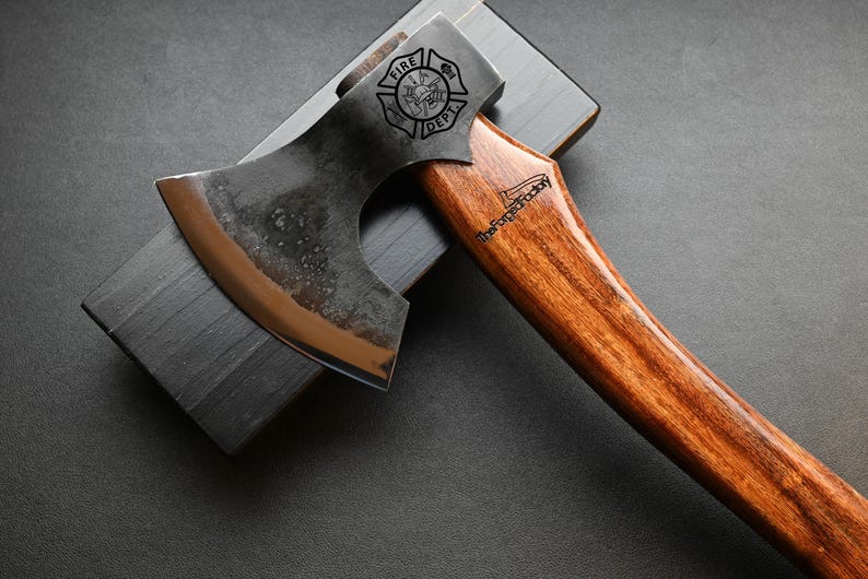 May include: Axe with a wooden handle and a metal head, resting on a dark gray sharpening stone. The axe head features the text "FIRE DEPT" and a logo. The handle has the text "TheForgedFactory".