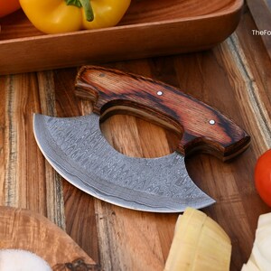 May include: A crescent-shaped ulu knife with a Damascus steel blade featuring a wavy pattern and a dark brown wood handle secured with two metal rivets. The blade is etched with the text TheForgedFactory and an axe icon.