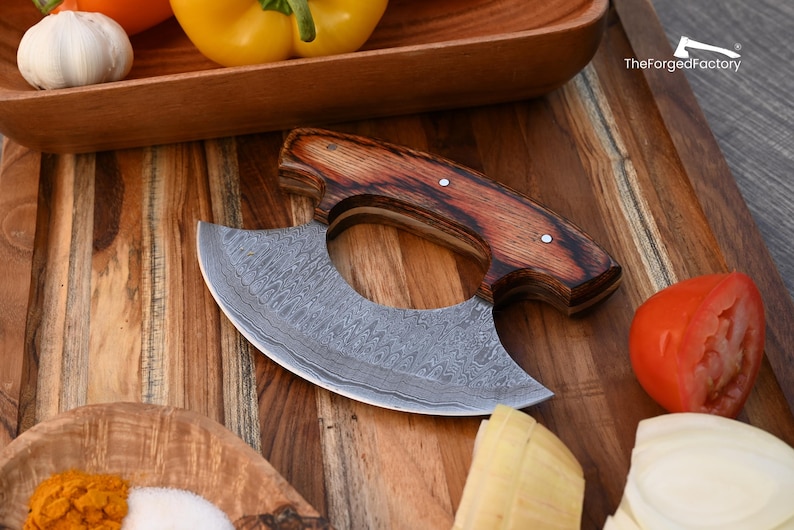 May include: A crescent-shaped ulu knife with a Damascus steel blade featuring a wavy pattern and a dark brown wood handle secured with two metal rivets. The blade is etched with the text TheForgedFactory and an axe icon.