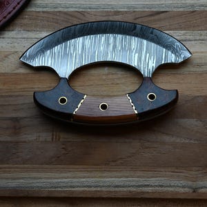 May include: A crescent-shaped ulu knife featuring a patterned Damascus steel blade and a handle constructed from three distinct segments of dark and light wood, secured with three brass rivets and decorative metal spacers.