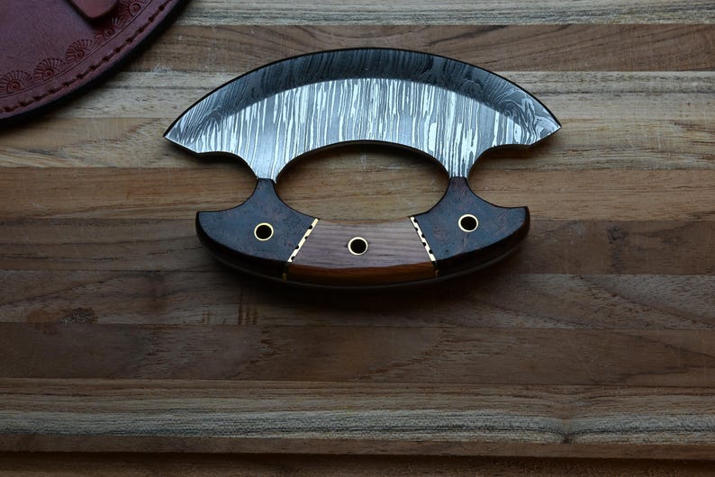 May include: A crescent-shaped ulu knife featuring a patterned Damascus steel blade and a handle constructed from three distinct segments of dark and light wood, secured with three brass rivets and decorative metal spacers.