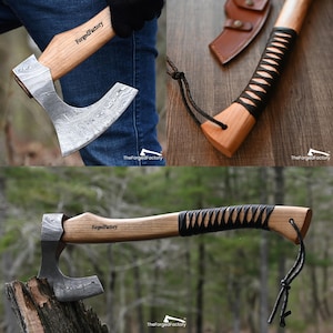 May include: A hand axe with a Damascus steel blade and a light wood handle featuring the text ForgedFactory. The handle includes a black leather wrap, a black cord lanyard, and a brown leather sheath. The axe head has a distinct wavy pattern.