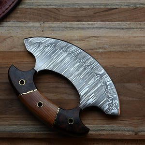 May include: A crescent-shaped ulu knife featuring a patterned Damascus steel blade and a handle constructed from two distinct types of dark and light brown wood, secured with three brass rivets.
