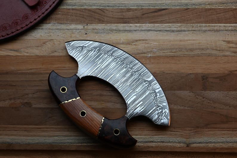 May include: A crescent-shaped ulu knife featuring a patterned Damascus steel blade and a handle constructed from two distinct types of dark and light brown wood, secured with three brass rivets.