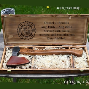 May include: A wooden box with a firefighter emblem and the text "Daniel J. Brooks Aug 1986 - Aug 2025". Inside is a wooden axe with a brown leather sheath. The axe is resting on a bed of straw.