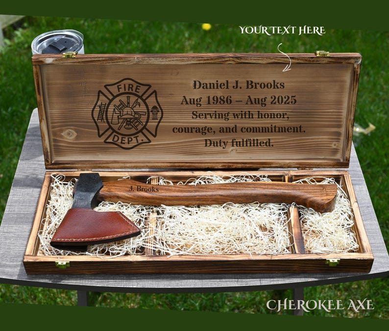 May include: A wooden box with a firefighter emblem and the text "Daniel J. Brooks Aug 1986 - Aug 2025". Inside is a wooden axe with a brown leather sheath. The axe is resting on a bed of straw.