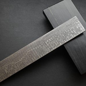 Peut inclure: Une lame métallique rectangulaire à motifs avec un motif ondulé repose sur une boîte en bois sombre. La lame a un aspect texturé unique, et la boîte est d'une couleur sombre unie. Le contraste entre les deux objets est saisissant.