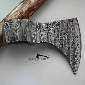 May include: A handcrafted axe head with a unique, patterned steel blade. The blade features a wavy, wood-grain-like design. The handle is a light brown color. The text "TheForgerFactory" is printed on the axe.