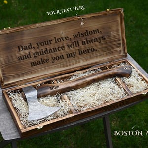 May include: A wooden box with a personalized axe inside, resting on a bed of straw. The box lid is open, displaying the engraved text "Dad, your love, wisdom, and guidance will always make you my hero." The axe handle is engraved with the name "Mike."