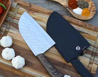 May include: A silver chef's knife with a wooden handle and a black leather sheath. The knife is lying on a wooden cutting board with three cloves of garlic.