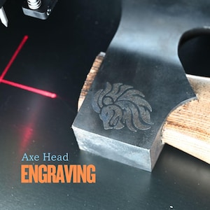 May include: Close-up of an axe head with a lion engraving. The axe head is dark gray with a wooden handle. The text "Axe Head Engraving" is displayed in orange and blue.