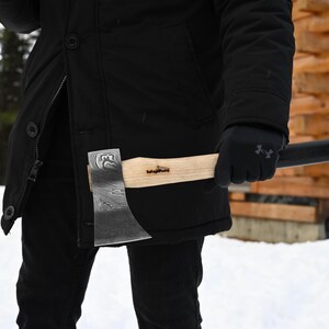 May include: A person wearing a black jacket and gloves holds a small axe with a wooden handle. The axe has a black blade with a patterned design and the text "Helle" on the handle.