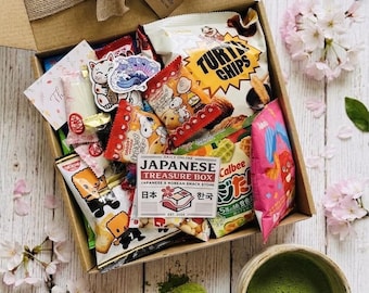 May include: A brown cardboard box filled with Japanese and Korean snacks, tied with a burlap bow. The box contains various colorful snack packages, stickers, and a small card. A cup of green matcha tea and a wooden spoon with matcha powder are next to the box.