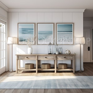 May include: A wooden console table with three drawers and two shelves. The table is decorated with three framed beach photos, two lamps, a vase with dried branches, and decorative items. The table is in front of a white wall with wood paneling.