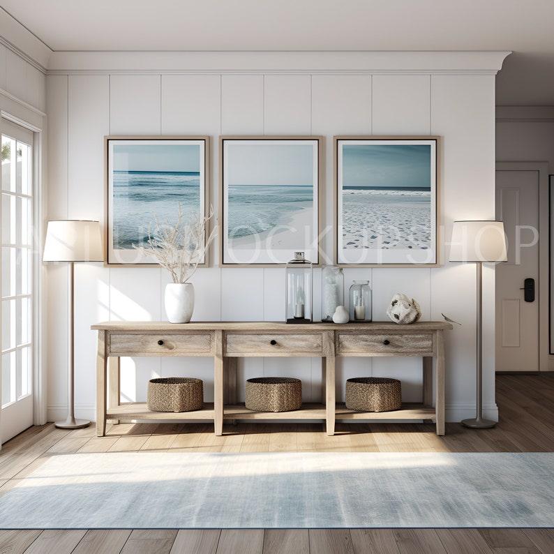 May include: A wooden console table with three drawers and two shelves. The table is decorated with three framed beach photos, two lamps, a vase with dried branches, and decorative items. The table is in front of a white wall with wood paneling.