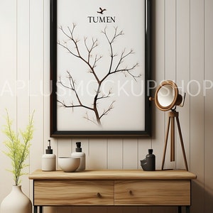 May include: A black framed print with a white background and a stylized tree branch design. The print has the word "TUMEN" in black text. The print is hanging on a white wall above a wooden console table with two drawers. The console table has black metal legs and is decorated with two white pump bottles, a white bowl, a black bottle, and a wooden tripod lamp with a round white shade.