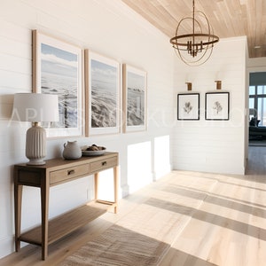 May include: A light wood console table with two drawers and a lower shelf. The table is decorated with a white ceramic pitcher, a bowl of decorative objects, and a white lamp with a beige shade. The table is in a hallway with white walls and a wooden floor. There are three framed pictures of seascapes on the wall above the table.