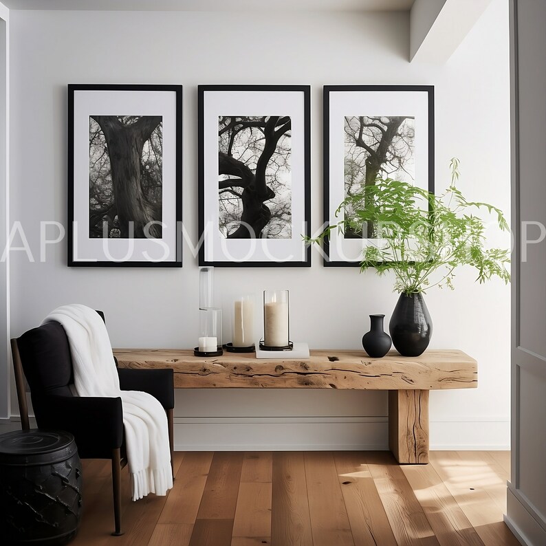 May include: Three black framed photographs of trees in a black and white color scheme hang on a white wall above a wooden bench with black candles and a vase of green plants. A black armchair with a white throw blanket sits to the left of the bench.