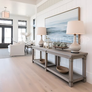 May include: A light wood console table with two shelves, two lamps with white shades, a framed abstract painting, and a decorative tray with succulents. The table is in a modern home with white walls and wood floors.