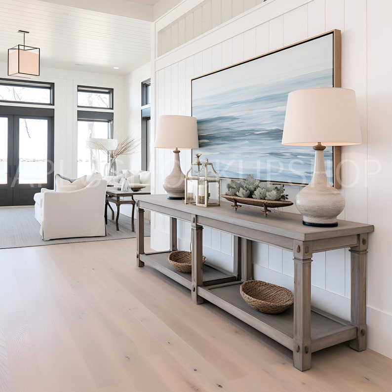 May include: A light wood console table with two shelves, two lamps with white shades, a framed abstract painting, and a decorative tray with succulents. The table is in a modern home with white walls and wood floors.