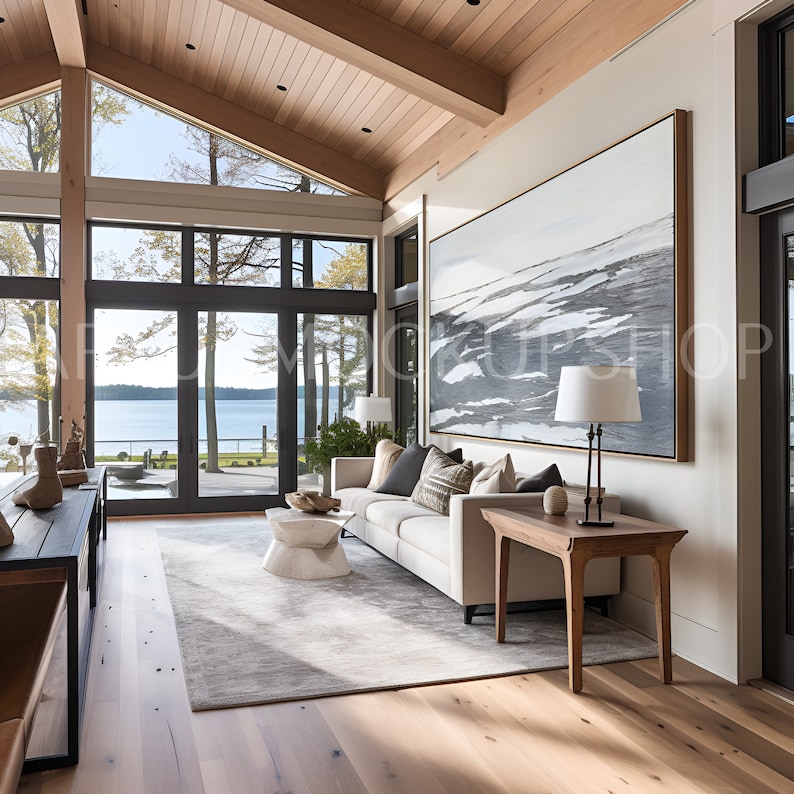 May include: A white sofa with pillows sits in a living room with large windows overlooking a lake. A wooden table with a lamp sits next to the sofa. A large abstract painting hangs on the wall above the table. The room has a wooden floor and a vaulted ceiling.