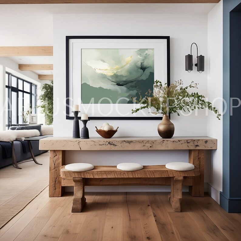 May include: A wooden console table with a rustic finish features a black framed abstract painting above it. The table has a light wood top and two benches with white cushions. A vase with green branches and a bowl of white decorative eggs are on the table.