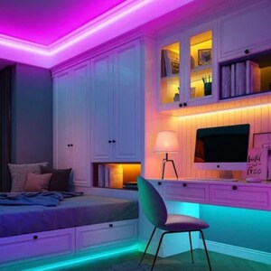 May include: A bedroom with a bed, a desk, and a bookshelf. The room is decorated with colorful LED lights. The lights are installed around the ceiling, under the bed, and under the desk. The desk has a computer monitor, a lamp, and a chair. The bookshelf is filled with books and has a few decorative items on top. The room is painted in a light color and has a modern design.