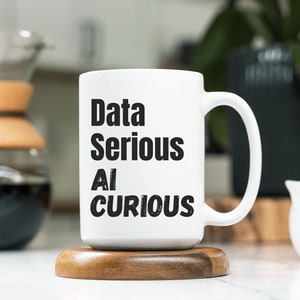 May include: White ceramic mug with black text that reads "Data Serious AI Curious". The mug is sitting on a wooden coaster.