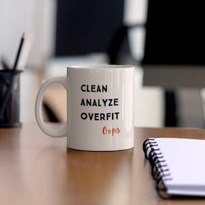 Op de afbeelding: Witte keramische koffiemok met de tekst "CLEAN ANALYZE OVERFIT Oops" in blauwe en oranje letters.
