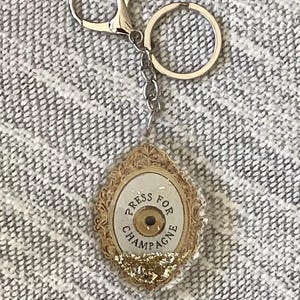 Könnte beinhalten: Ein Schlüsselanhänger zum Thema Champagner mit einem goldfarbenen ovalen Anhänger. Der Anhänger trägt die Aufschrift "PRESS FOR CHAMPAGNE" in einem kreisförmigen Design. Der Schlüsselanhänger enthält einen silbernen Verschluss und einen Ring.