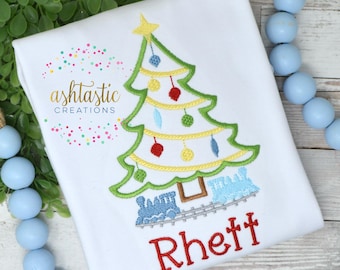 May include: A white onesie with an embroidered Christmas tree with ornaments and a train underneath. The name "Rhett" is embroidered in red below the train.