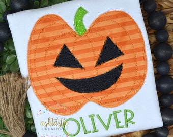 May include: A white t-shirt with an orange and black pumpkin applique. The pumpkin has a stitched-on smile and eyes. The text "OLIVER" is stitched in green below the pumpkin.