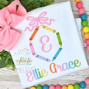 May include: A white t-shirt with a pink bow and a hexagon made of colorful crayons surrounding the letter "E". The name "Ellie Grace" is embroidered in rainbow colors below the hexagon.