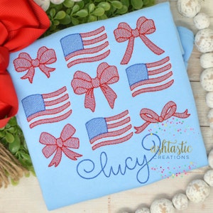 May include: Light blue shirt embroidered with red bows and American flags. The name "Lucy" is embroidered in a cursive font. The shirt also has the text "Ashtastic Creations" in a script font.