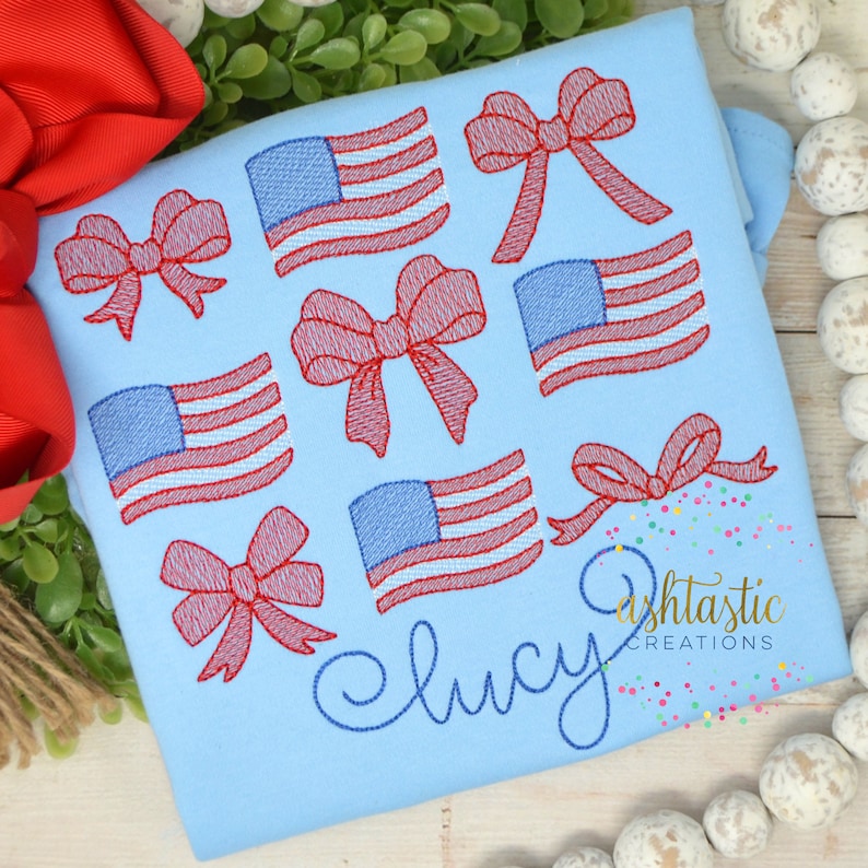 May include: Light blue shirt embroidered with red bows and American flags. The name "Lucy" is embroidered in a cursive font. The shirt also has the text "Ashtastic Creations" in a script font.