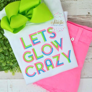 May include: White t-shirt with the text "Let's Glow Crazy" in neon green, pink, and blue outlined letters. The shirt is folded and displayed with a pink pair of shorts.