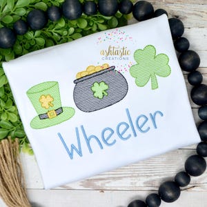May include: A white onesie with a green leprechaun hat, a pot of gold, and a shamrock. The name "Wheeler" is embroidered in blue below the design.