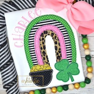 May include: A white t-shirt with a pink, black, and green rainbow design. The rainbow has a leopard print inside. Underneath the rainbow is a black pot with gold coins and a green shamrock. The shirt is personalized with the name "Charlie".