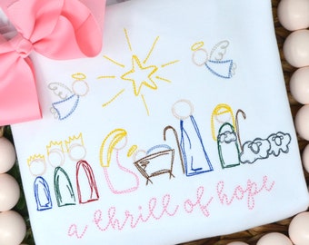 May include: A white onesie with a pink bow on the top left corner. The onesie has an embroidered image of the nativity scene with a star and angels. The text "a thrill of hope" is embroidered in pink below the image.