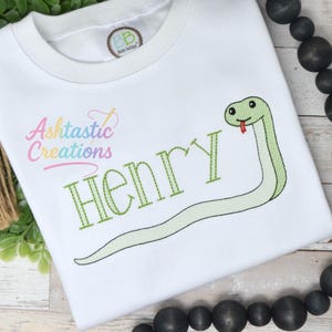 May include: White t-shirt with a green embroidered snake and the name "Henry" in green stitching. The shirt features the "Ashtastic Creations" logo in pink and the "BB" logo on the collar.