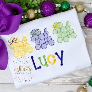 May include: A white shirt with three embroidered Mardi Gras beads in the shape of dogs. The dogs are yellow, purple, and green. The name "Lucy" is embroidered below the dogs in yellow, green, and purple letters. The shirt has a confetti pattern in the bottom left corner.