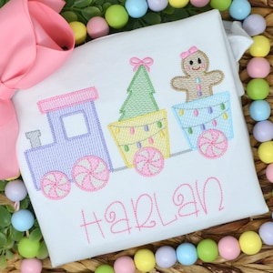 May include: A white embroidered shirt with a pink, purple, and green train carrying a gingerbread person, a Christmas tree, and candy canes. The name "HARLAN" is embroidered below the train.