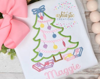 May include: A white onesie with a green embroidered Christmas tree with pink, blue, and yellow ornaments. The tree has a blue bow at the top and is surrounded by pink and purple gift boxes. The name "Maggie" is embroidered in pink cursive below the tree.