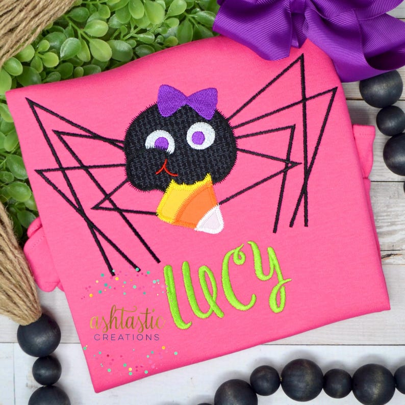 May include: Pink t-shirt with an embroidered black spider holding a candy corn. The spider has a purple bow and blue eyes. The name "Lucy" is embroidered in green below the spider.